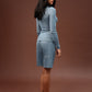 A woman stands barefoot in profile wearing a light blue matching lounge set consisting of a long-sleeve top and shorts, against a solid dark brown studio background.