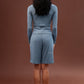 A woman stands with her back to the camera wearing a matching dusty blue long-sleeve top and shorts set against a solid brown studio backdrop.