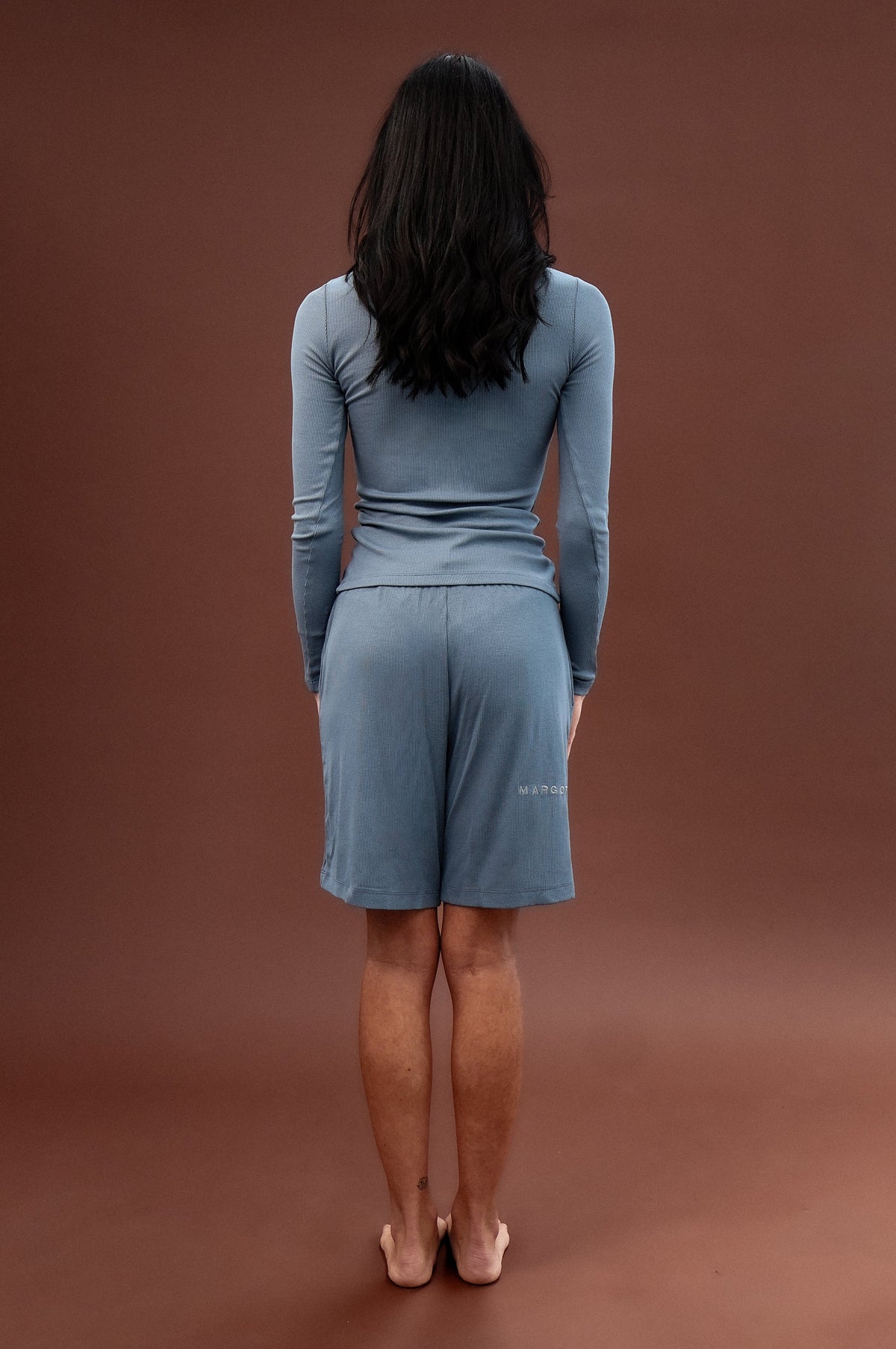 A woman stands with her back to the camera wearing a matching dusty blue long-sleeve top and shorts set against a solid brown studio backdrop.