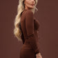 A blonde woman posing in a side profile wearing a brown long-sleeve knit dress against a solid dark brown studio background.