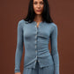 A woman modeling a matching dusty blue ribbed knit loungewear set, featuring a long-sleeve button-front top and coordinating pants, set against a solid dark brown studio background.