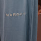 Close-up of a person wearing slate blue ribbed lounge pants with the brand name 'MARGOT' embroidered in beige on the left thigh, paired with a matching long-sleeve top against a solid brown background.