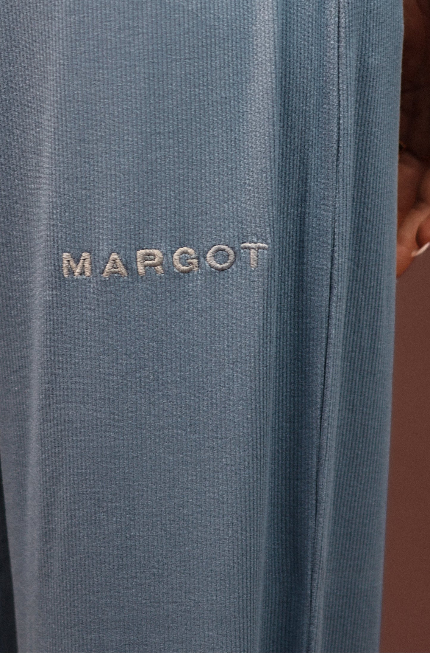 Close-up of a person wearing slate blue ribbed lounge pants with the brand name 'MARGOT' embroidered in beige on the left thigh, paired with a matching long-sleeve top against a solid brown background.