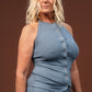A woman with blonde hair wearing a slate blue sleeveless button-front top and matching pants, standing against a solid dark brown studio background.