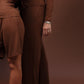 Two women modeling matching brown long-sleeve lounge sets in a studio setting with a solid brown backdrop. The woman in front wears a matching short set and a patterned headscarf, while the woman behind wears a matching long pants set.