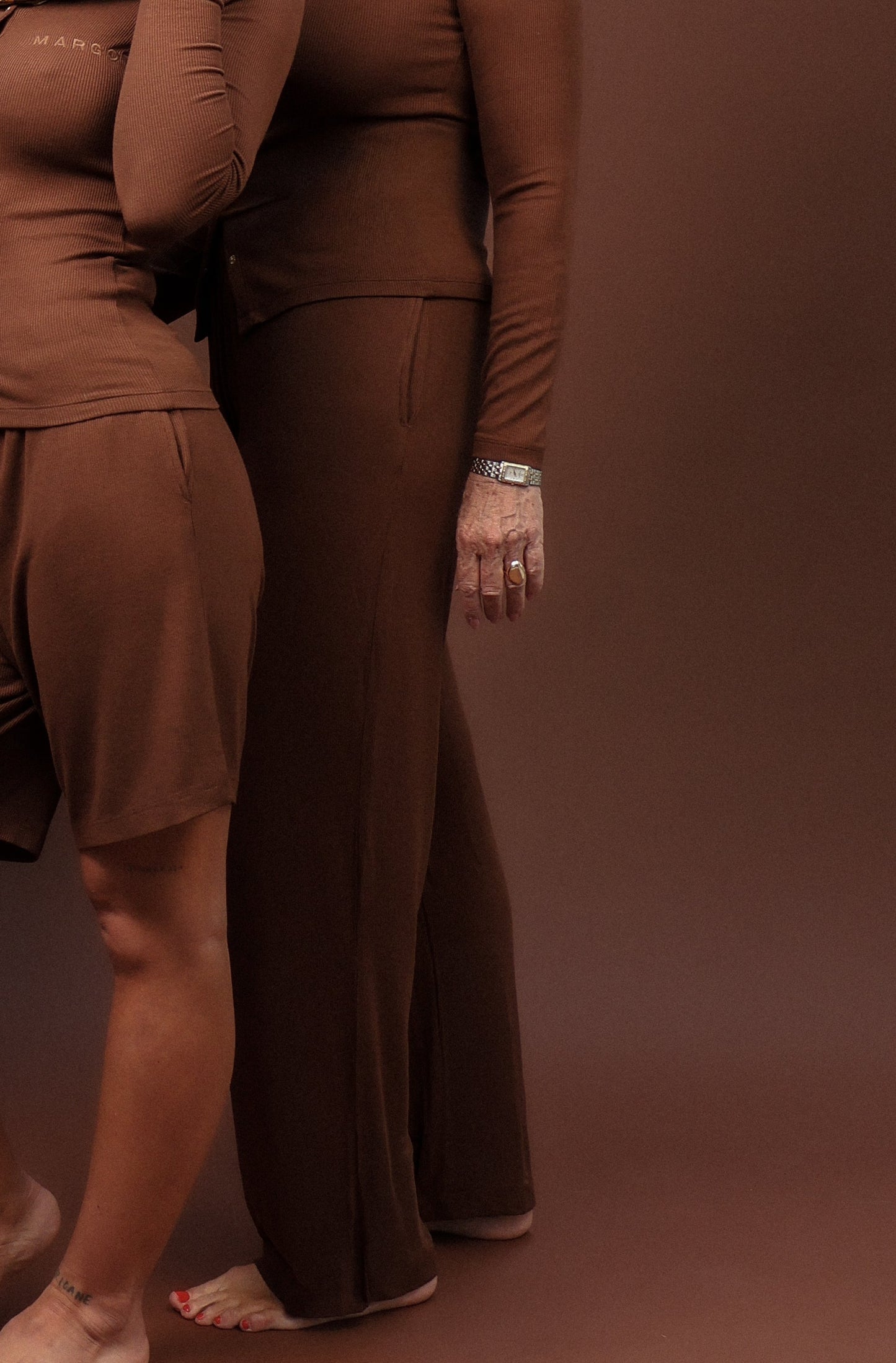 Two women modeling matching brown long-sleeve lounge sets in a studio setting with a solid brown backdrop. The woman in front wears a matching short set and a patterned headscarf, while the woman behind wears a matching long pants set.