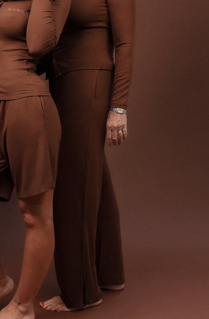 Two women modeling matching brown long-sleeve lounge sets in a studio setting with a solid brown backdrop. The woman in front wears a matching short set and a patterned headscarf, while the woman behind wears a matching long pants set.
