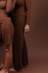 Two women modeling matching brown long-sleeve lounge sets in a studio setting with a solid brown backdrop. The woman in front wears a matching short set and a patterned headscarf, while the woman behind wears a matching long pants set.