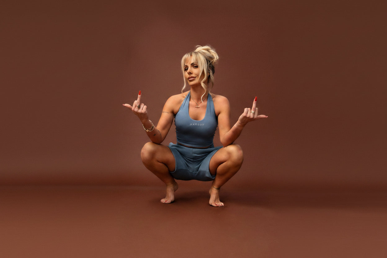 A blonde woman in a blue athletic matching set crouches in a studio setting against a solid brown backdrop, holding up two fingers on each hand.