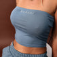 A close-up of a woman wearing a dusty blue ribbed strapless crop top with 'MARGOT' printed in white on the chest, paired with matching blue ruched bottoms.