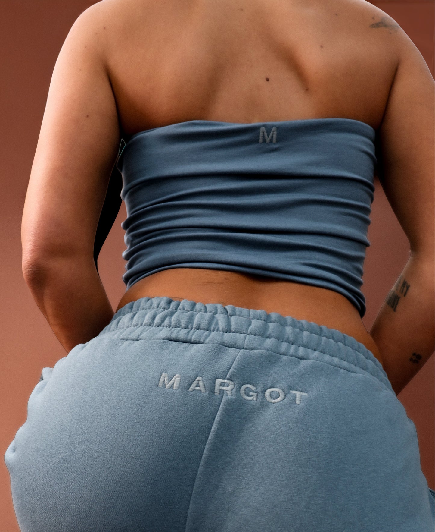 Rear view of a person wearing a dusty blue strapless tube top and matching sweatpants with 'MARGOT' embroidered on the back pocket, against a warm brown background.