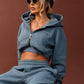 A woman wearing a matching dusty blue lounge set consisting of a cropped zip-up hoodie and wide-leg sweatpants, sitting on a small black stool against a solid dark brown studio backdrop.