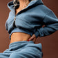 A model wearing a matching dusty blue cropped zip-up hoodie and sweatpants set, seated on a black stool against a solid brown studio background.