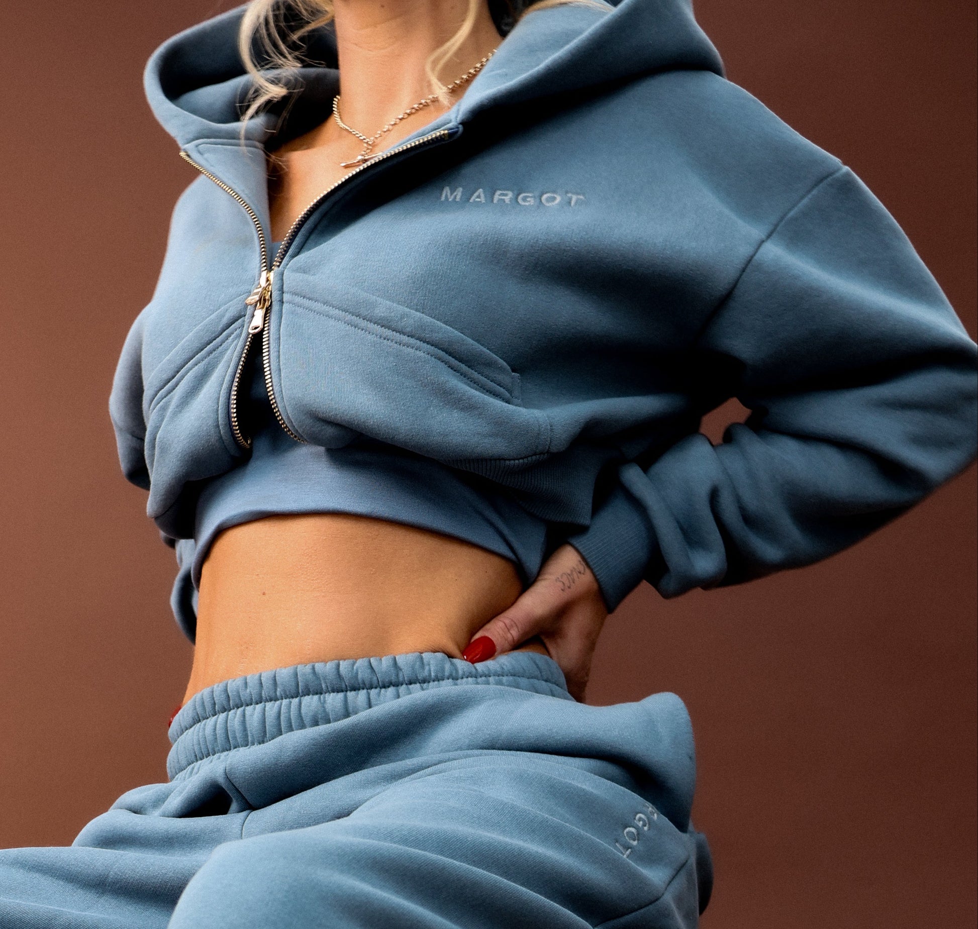 A model wearing a matching dusty blue cropped zip-up hoodie and sweatpants set, seated on a black stool against a solid brown studio background.