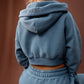 A person shown from behind wearing a dusty blue cropped hoodie and matching sweatpants, both with the brand name 'MARGOT' embroidered on them. The background is a solid warm brown wall.