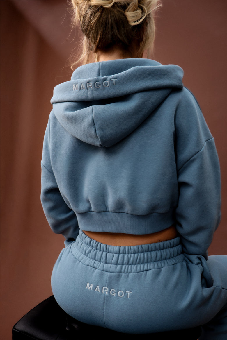 A person shown from behind wearing a dusty blue cropped hoodie and matching sweatpants, both with the brand name 'MARGOT' embroidered on them. The background is a solid warm brown wall.