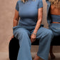 Three women modeling a matching blue loungewear set in a studio setting with a beige textured backdrop. One woman is seated on a black bench, showing the front of the matching set, while the other two show the back of the matching hooded set.