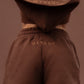 A model shown from behind wearing a matching brown cropped hoodie and shorts set with 'MARGOT' embroidered on the back of each piece, set against a solid brown background.