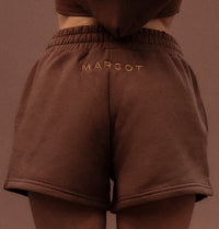 A model shown from behind wearing a matching brown cropped hoodie and shorts set with 'MARGOT' embroidered on the back of each piece, set against a solid brown background.