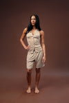 A woman modeling a light beige sleeveless button-front romper with matching shorts, standing against a solid dark brown studio backdrop.