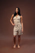 A woman modeling a light beige sleeveless button-front romper with matching shorts, standing against a solid dark brown studio backdrop.